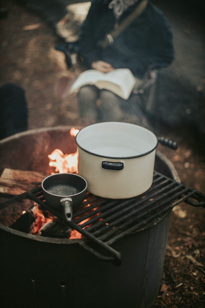 Close-up of a campfire cooking setup with cast iron pots on a grill, while a person reads comfortably nearby, evoking cozy backyard patio evenings.