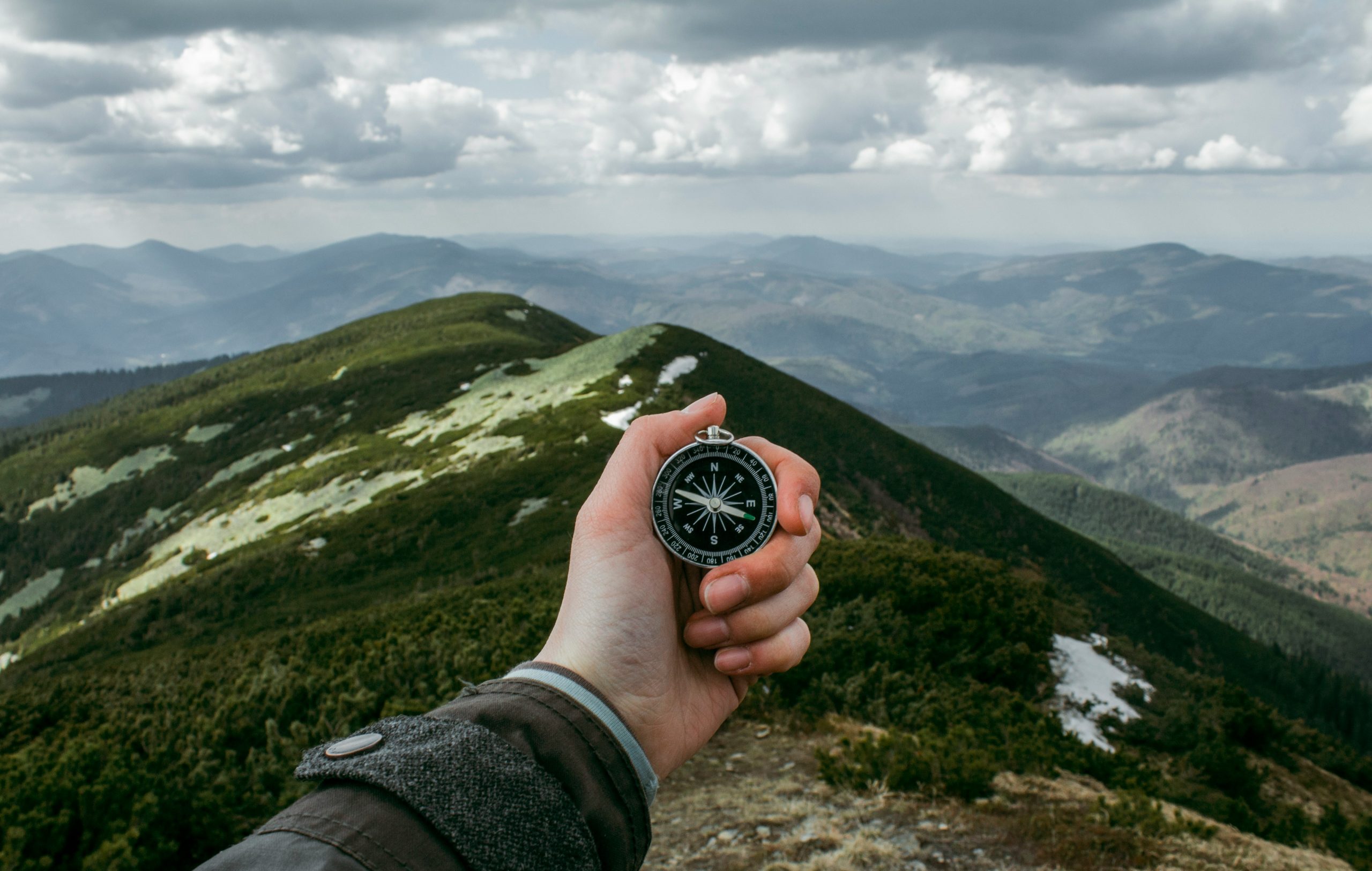 A hand holding a black compass with white dials against a backdrop of misty, snow-dusted mountain peaks - symbolizing navigation and durable outdoor exploration