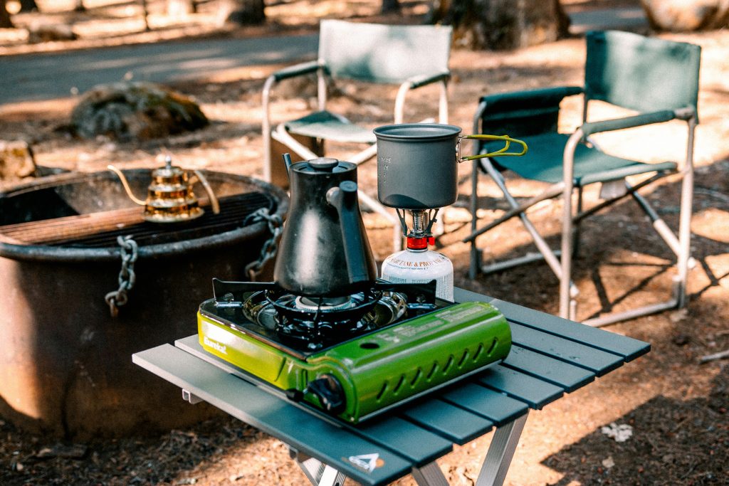 A complete portable camping kitchen setup with a green stove, kettle, and pot on a folding table in the woods, ideal for durable outdoor cooking and patio gear.